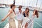 A group of young handsome models is standing on the bow of the yacht and having a good time while riding through the dock on the