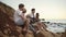 Group of young hipster friends sitting on the rocks by the seashore and playing guitar and singing songs. Slowmotion