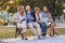 Group of young  people laughing together in park, sitting on bench, holding their cell phones, laptop, tablet. sharing  good news