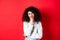 Grumpy young woman with curly hair, looking annoyed or bored at empty space, standing pensive and sad on red background