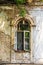 Grungy arched casement window in wall with plaster peeling off exposing bricks underneath - striped curtains and plants in open