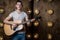 Guitarist, music. A young man plays an acoustic guitar on a background with lights behind him. Horizontal frame