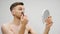 Guy applies himself makeup. Young handsome man applying makeup on face. Bearded gay making makeup while looking on mirror