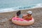 Guy in a cap sits in an inflated circle in the form of a donut with pink icing on the beach and works in a laptop online. The