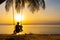 The guy enjoys the sunset riding on a swing on the ptropical beach. Silhouettes of a guy on a swing hanging on a palm tree,