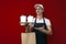Guy a food delivery uniform in uniform gives coffee and a package on a red background, delivery service worker