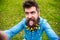 Guy with lesser celandine flowers in beard taking selfie photo. Hipster with cheerful grimace with tongue on face taking