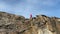 A guy in a red jacket walks along the edge of a rocky mountain.