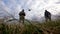 A guy with a remote control and a girl are standing on the ground. A drone flies over them