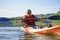 The guy is sailing on a red kayak, wearing a life jacket with glasses and a cap. Holds a paddle in his hand