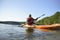 The guy is sailing on a red kayak, wearing a life jacket with glasses and a cap. Holds a paddle in his hand