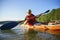 The guy is sailing on a red kayak, wearing a life jacket with glasses and a cap. Holds a paddle in his hand