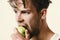 Guy with serious face isolated on light grey background, close up. Man with green apple in his hand bites it. Athlete