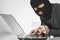 Hacker in balaclava holding a card in left hand and typing something with right hand on laptop keyboard