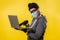 A hacker in a medical mask is holding a laptop. Yellow background. The concept of cybercrime growth in the context of the