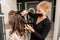 A hairdresser in a black mask prepares a woman, makes a hairstyle with curls using a curling iron. Long light brown