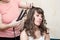 Hairdresser curling hair strand of model with flat iron, domestic room, two girls