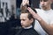 The hairdresser makes a fashionable pretty hairstyle for the boy in a modern barbershop.