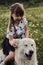 Half breed white Swiss shepherd in chamomile field lies at smiles. Cute little Caucasian girl sits on big white dog and smiles.