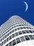 Half a moon over a building skyscraper, shining in bright blue sky. Below the crescent of quarter moon over a tall