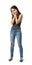 Half-turn view of young sad woman in gray sleeveless top and blue jeans standing with hands at face isolated on white