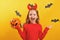 Halloween celebration. Little girl in a devil costume holds a bucket of pumpkin on a yellow background