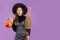 Halloween theme, young asian girl in witch costume holding broom and carrying orange pumpkin bucket posing on purple color