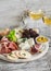 Ham, cheese, grapes, figs, nuts, bread ciabatta, cracker, jam on white wooden board and two glasses of white wine on bright wooden