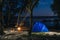 Hammok and girl is sitting near bonfire. Blue Camping Tent Illuminated Inside. Night Hours Campsite. Recreation and Outdoor. Lake
