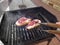 A hand with barbecue tongs picking up a beef steak that is being grilled over embers, two veal chops on a barbecue