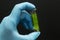 A hand in a blue medical glove holds a glass test tube with a green leaf inside