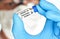 Hand in blue medical gloves holding small vial with label own design, dummy barcode Covid 19 vaccine, blurred face in cotton