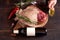 Hand, with a container of olive oil, next to a piece of juicy, seasoned rib eye steak wrapped in paper and burlap