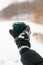 Hand in cozy glove holding reusable cup of tea or coffee on background of snowy lake in winter. Hiking and traveling in cold