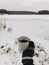 Hand in cozy glove holding warm cup of tea on background of snowy lake in winter. Hiking and traveling in cold winter season. Warm