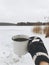 Hand in cozy glove holding warm cup of tea on background of snowy lake in winter. Hiking and traveling in cold winter season. Warm