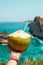 Hand with fresh coconut, ocean and cliffs tropical landscape. Vacation mood. Bali, Indonesia