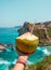 Hand with fresh coconut, ocean and cliffs tropical landscape. Vacation mood. Bali, Indonesia