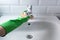 Hand in green rubber gloves clean sink in domestic bathroom yellow sponge and liquid foam detergent. Concept housework