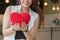 Hand of happily smiling beautiful young woman standing and holding a small red heart pillow in front of cafe. Valentine