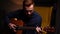 Hand held shot of talented young guitarist man playing guitar sitting on armchair in dark living room at home studio.