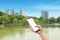Hand holding black smartphone with blurred cityscape park view and blue sky in background.