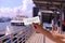 Hand holding blank mockup for one-fold ticket design, at a pier. Cruise ferry and incidental people in the background, defocused.
