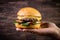 Hand holding a craft beef burger with cheese, rocket leafs and french fries on rustic background