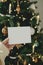 Hand holding empty greeting card on background of stylish decorated christmas tree with golden lights. Christmas card mock up.