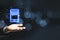 Hand holding glowing smartphone icon hologram on dark bokeh background with mockup place. Smart technology concept