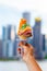 Hand holding ice cream cone with several layers of different colors with blurred city in the background