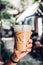 Hand holding iced latte with straw in plastic cup at garden blur cafe restaurant in hot sunny day.coffee shop,cafe leisure