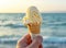 A hand holding a melting vanilla ice cream cone against a serene beach.