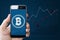Hand holding mobile smartphone with B symbol of Bitcoin, internet banking and block chain on screen and raising graph background